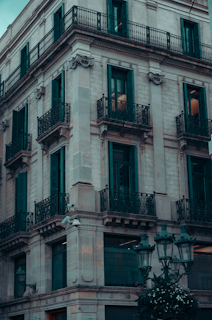 A classical European-style building with ornate wrought iron balconies and large windows. The architecture features detailed stonework and a corner viewpoint. Street lamps and a hint of greenery at the base add to the urban setting.