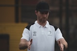 A man wearing a black cap and a white polo shirt with black shoulder accents gestures with his hands. The shirt has logos including 'Umbro' and 'Alliance', indicating possible sports or coaching affiliation. The background appears blurred, suggesting an outdoor setting.