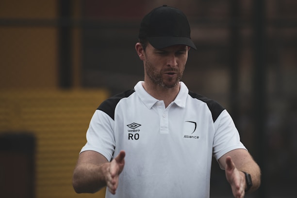 A man wearing a black cap and a white polo shirt with black shoulder accents gestures with his hands. The shirt has logos including 'Umbro' and 'Alliance', indicating possible sports or coaching affiliation. The background appears blurred, suggesting an outdoor setting.
