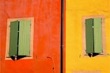 A vibrant facade featuring two sections of a building wall. The left section is painted in a bold orange-red color, and the right section in a bright yellow. Each section contains a window with closed green shutters, framed by a neutral-colored border. A vertical black drainpipe divides the two sections.