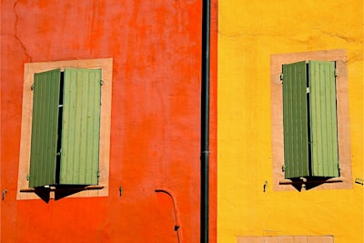 A vibrant facade featuring two sections of a building wall. The left section is painted in a bold orange-red color, and the right section in a bright yellow. Each section contains a window with closed green shutters, framed by a neutral-colored border. A vertical black drainpipe divides the two sections.