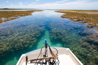 A small boat is navigating through clear, shallow waters with visible coral reefs below. The water is a vibrant turquoise contrasting with the sandy and rocky shores. The horizon stretches out under a blue sky with scattered clouds.