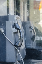 An old payphone with a black handset and a silver, coiled cord. The surrounding area appears to be enclosed in glass, with some streaks and marks visible on the surface.
