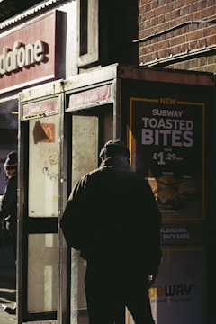 A person in dark clothing stands near an old, weathered phone booth. The scene is set on a street corner, where sunlight casts deep shadows. Advertisements for fast food are visible, and the words 'Subway Toasted Bites' are prominently displayed. The backdrop includes a brick wall and a partial view of a Vodafone store sign.