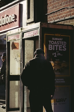 A person in dark clothing stands near an old, weathered phone booth. The scene is set on a street corner, where sunlight casts deep shadows. Advertisements for fast food are visible, and the words 'Subway Toasted Bites' are prominently displayed. The backdrop includes a brick wall and a partial view of a Vodafone store sign.