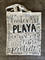 A canvas tote bag with various words in decorative black lettering, including 'PLAYA' in bold. The bag features floral and vine designs in muted gray tones.