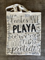 A canvas tote bag with various words in decorative black lettering, including 'PLAYA' in bold. The bag features floral and vine designs in muted gray tones.