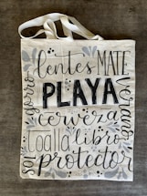 A canvas tote bag with various words in decorative black lettering, including 'PLAYA' in bold. The bag features floral and vine designs in muted gray tones.
