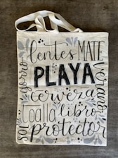 A canvas tote bag with various words in decorative black lettering, including 'PLAYA' in bold. The bag features floral and vine designs in muted gray tones.