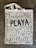 A canvas tote bag with various words in decorative black lettering, including 'PLAYA' in bold. The bag features floral and vine designs in muted gray tones.