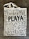 A canvas tote bag with various words in decorative black lettering, including 'PLAYA' in bold. The bag features floral and vine designs in muted gray tones.