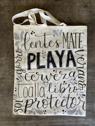 A canvas tote bag with various words in decorative black lettering, including 'PLAYA' in bold. The bag features floral and vine designs in muted gray tones.