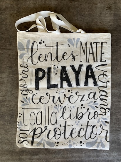 A canvas tote bag with various words in decorative black lettering, including 'PLAYA' in bold. The bag features floral and vine designs in muted gray tones.
