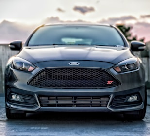 A black Ford car is parked, featuring a sleek design with a prominent front grille and headlights. The emblem is visible in the center of the grille. Soft lighting indicates a dusk or dawn setting, with blurred background elements.