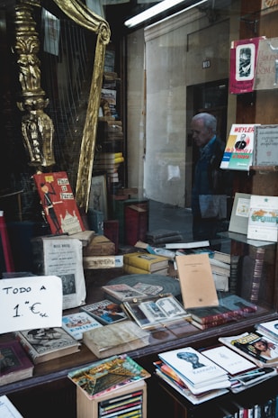 A storefront window displays a collection of old books, magazines, and a sign indicating everything is priced at 1 Euro. A golden harp and various decorations are visible among the items. Outside the window, a person is seen walking past.