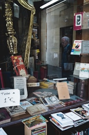 A storefront window displays a collection of old books, magazines, and a sign indicating everything is priced at 1 Euro. A golden harp and various decorations are visible among the items. Outside the window, a person is seen walking past.