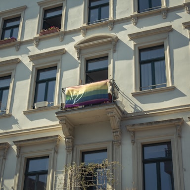 A building facade with multiple windows featuring classic architectural details. A rainbow flag is draped over a small balcony railing, which adds a vibrant touch to the otherwise neutral colors of the building.