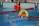 A swimmer in a pool is seen mid-action holding an orange and yellow flotation device with the text 'RLSS UK Speeds'. The swimmer is wearing a white swim cap and competing in a lane marked by red and blue lane dividers. A lifeguard or coach's legs and an orange buoy are visible at the poolside.