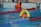A swimmer in a pool is seen mid-action holding an orange and yellow flotation device with the text 'RLSS UK Speeds'. The swimmer is wearing a white swim cap and competing in a lane marked by red and blue lane dividers. A lifeguard or coach's legs and an orange buoy are visible at the poolside.