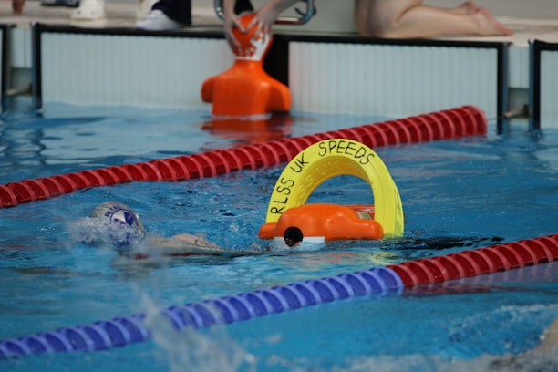 A swimmer in a pool is seen mid-action holding an orange and yellow flotation device with the text 'RLSS UK Speeds'. The swimmer is wearing a white swim cap and competing in a lane marked by red and blue lane dividers. A lifeguard or coach's legs and an orange buoy are visible at the poolside.