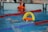 A swimmer in a pool is seen mid-action holding an orange and yellow flotation device with the text 'RLSS UK Speeds'. The swimmer is wearing a white swim cap and competing in a lane marked by red and blue lane dividers. A lifeguard or coach's legs and an orange buoy are visible at the poolside.