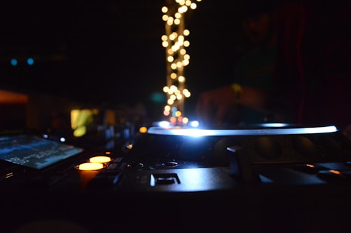 A dimly lit DJ setup with glowing equipment, surrounded by colorful lights and a blurred figure in the background.