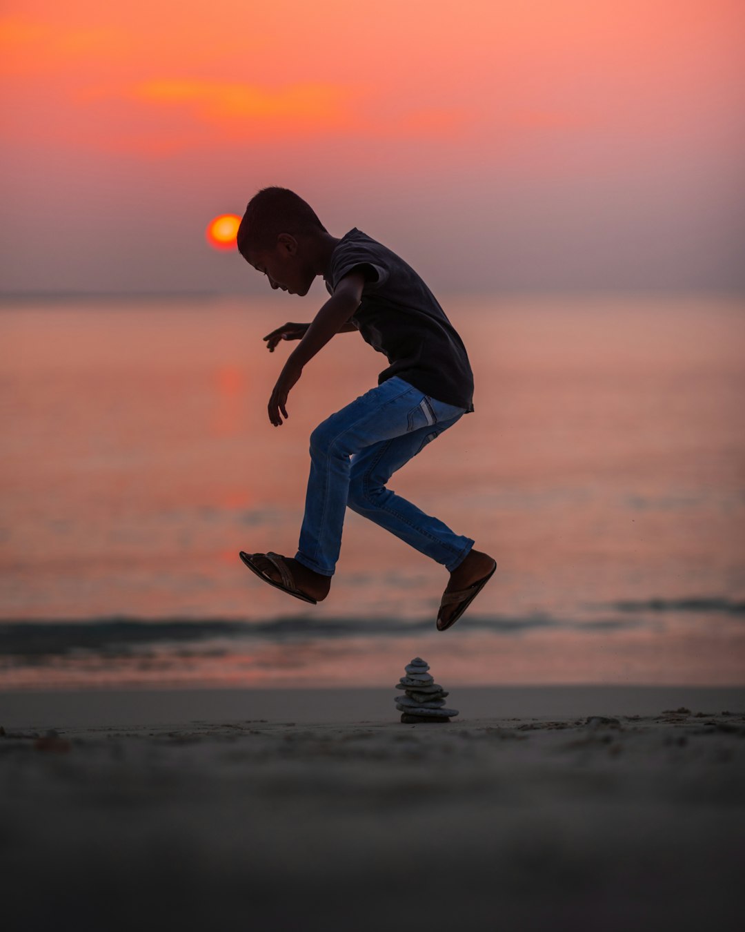 boy jumping near stack of stones on seashore during golden hour