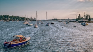 A scenic harbor filled with numerous sailboats and yachts gently floating on the water. In the forefront, a small, blue motorboat with a wooden deck and an American flag at the stern floats in the shimmering sunlight. The background is lined with lush green trees and distant buildings on the shore under a partly cloudy sky.