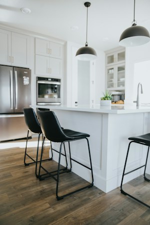 A modern kitchen with sleek design elements, featuring two black pendant lights hanging over a white kitchen island. Black leather bar stools with metal frames are placed around the island. The cabinetry is light-colored with modern stainless steel appliances integrated into the design. The flooring is wooden, adding warmth to the contemporary style.