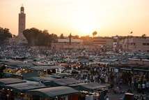 A bustling market scene during sunset, with numerous stalls covered by green tarps. Many people are present, both shopping and socializing. In the background, a tall minaret stands out against the setting sun, casting a warm glow over the buildings.