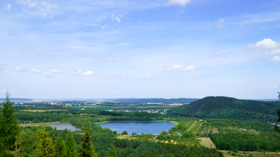 The image presents a panoramic view of a lush, green landscape with a large lake nestled amidst the verdant surroundings. The lake is surrounded by an array of trees and shrubs, creating a serene and tranquil atmosphere. In the distance, there are several buildings scattered across the landscape, adding depth to the scene. The sky above is a clear blue, dotted with fluffy white clouds that add to the overall beauty of the image.