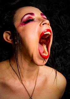A person with bold makeup, featuring bright red lipstick and vivid pink eyeshadow, dramatically opens their mouth wide. Eyebrows are styled in a prominent fashion, complemented by dark eyeliner. A small piercing is visible on the lower lip, and a nostril ring is also present. The person is wearing a necklace, and their hair appears to be styled upwards. The background is a textured dark pattern, adding depth to the composition.