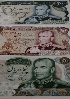 Three different Iranian banknotes featuring detailed engravings of a man's portrait dressed in military attire. The notes are adorned with intricate floral and scrollwork patterns, accompanied by Persian script.