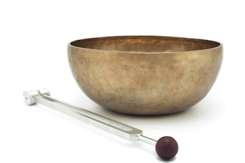 A large brass-colored singing bowl set on a white surface is accompanied by a metal mallet with a brown rubber head.