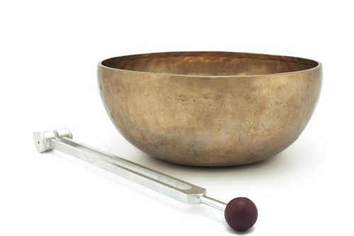 A large brass-colored singing bowl set on a white surface is accompanied by a metal mallet with a brown rubber head.
