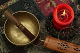 A brass singing bowl with a wooden mallet rests on an intricately patterned textile. Nearby, a red candle burns with a visible flame, casting a warm glow. An ornate incense box releases a trail of smoke, suggesting an atmosphere of tranquility and meditation.