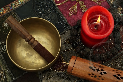 A brass singing bowl with a wooden mallet rests on an intricately patterned textile. Nearby, a red candle burns with a visible flame, casting a warm glow. An ornate incense box releases a trail of smoke, suggesting an atmosphere of tranquility and meditation.