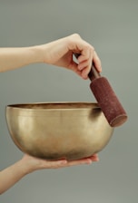 A hand holds a large, golden singing bowl, with another hand gently holding a mallet over the bowl's rim. The background is a soft, muted green, creating a calm atmosphere.