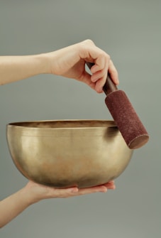 A hand holds a large, golden singing bowl, with another hand gently holding a mallet over the bowl's rim. The background is a soft, muted green, creating a calm atmosphere.