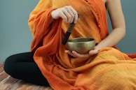 A person is dressed in an orange robe and holding a small brass bowl and a wooden mallet. The individual appears to be sitting cross-legged on a mat, and the background is a solid blue-gray color.
