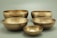 Five metallic singing bowls of varying sizes are displayed against a soft green background. The bowls have a brass-like appearance, featuring a hammered texture on their surfaces.
