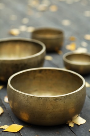 Several metallic singing bowls of varying sizes are arranged on a dark wooden surface scattered with autumn leaves. The focus is on the largest bowl in the foreground, with the rest softly blurred in the background.