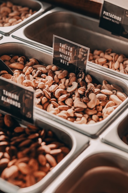 A variety of nuts displayed in metal containers, each labeled with signs indicating the type of nuts. The focus is on a bin containing salted deluxe mixed nuts. In the background, other bins contain pistachios and Tahitian almonds. The metallic finish of the containers contrasts with the earthy tones of the nuts.