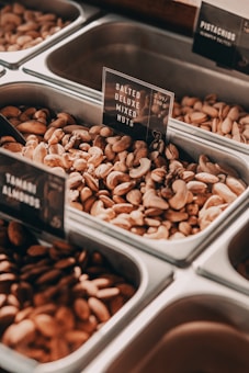 A variety of nuts displayed in metal containers, each labeled with signs indicating the type of nuts. The focus is on a bin containing salted deluxe mixed nuts. In the background, other bins contain pistachios and Tahitian almonds. The metallic finish of the containers contrasts with the earthy tones of the nuts.