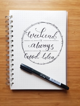 A spiral-bound notebook with dotted pages is open to a page featuring handwritten calligraphy that reads 'Weekend is always a good idea.' A black marker is placed diagonally on the notebook. The backdrop is a wooden surface.