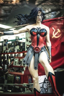 A detailed and realistic statue of a woman in a superhero costume is displayed inside a store. The costume is predominantly red and black with a star emblem on her chest and head. She is holding a Soviet Union flag with a hammer and sickle symbol. The background features numerous comic books and posters, one of which depicts the Joker character.