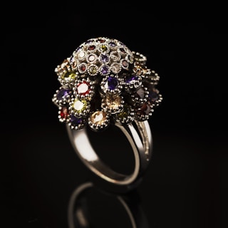 A luxurious and ornate ring featuring numerous small, multifaceted gemstones in various colors is elegantly showcased. The ring is crafted from shiny metal and is set against a stark black background, highlighting its intricate design and vibrant gemstones.