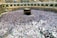 A large crowd of people gathered around the Kaaba in the center of the Grand Mosque in Mecca, wearing white garments, performing a religious ritual. The mosque is surrounded by arched colonnades and multi-level balconies, extending into the background.