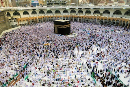 A large crowd of people gathered around the Kaaba in the center of the Grand Mosque in Mecca, wearing white garments, performing a religious ritual. The mosque is surrounded by arched colonnades and multi-level balconies, extending into the background.