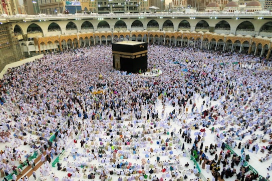 A large crowd of people gathered around the Kaaba in the center of the Grand Mosque in Mecca, wearing white garments, performing a religious ritual. The mosque is surrounded by arched colonnades and multi-level balconies, extending into the background.